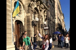 [ai] A woman with long brown hair poses with a smile in front of a historical building featuring a Ukrainian flag. Other people walk past, engaged in various activities, in a lively urban setting.
